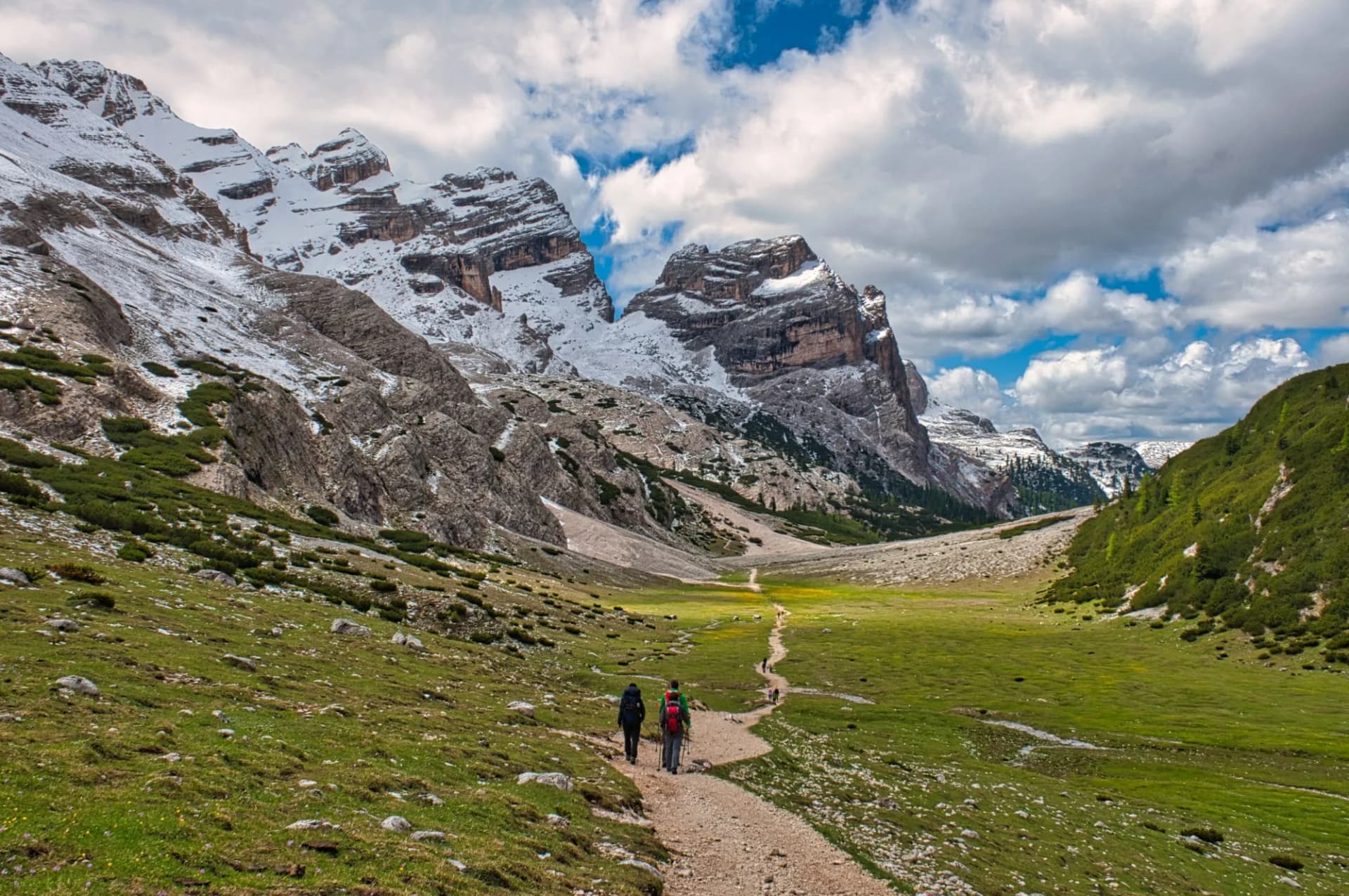 Hikers on dirt path through green valley toward snow-capped mountains under cloudy sky