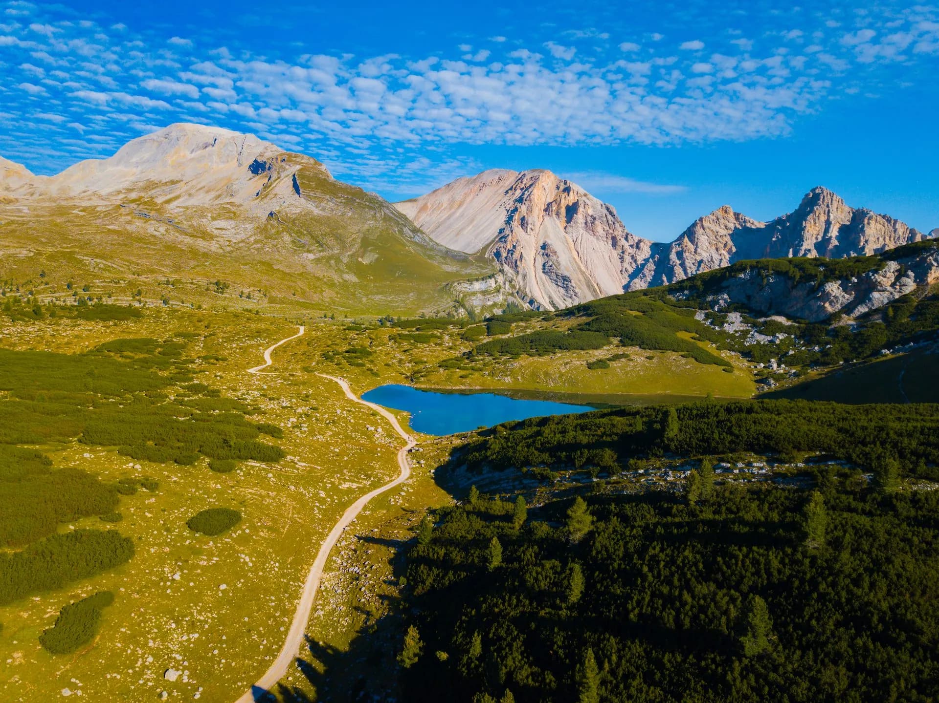 Alpine lake with bright blue water, dirt path, and rugged mountains under blue sky.