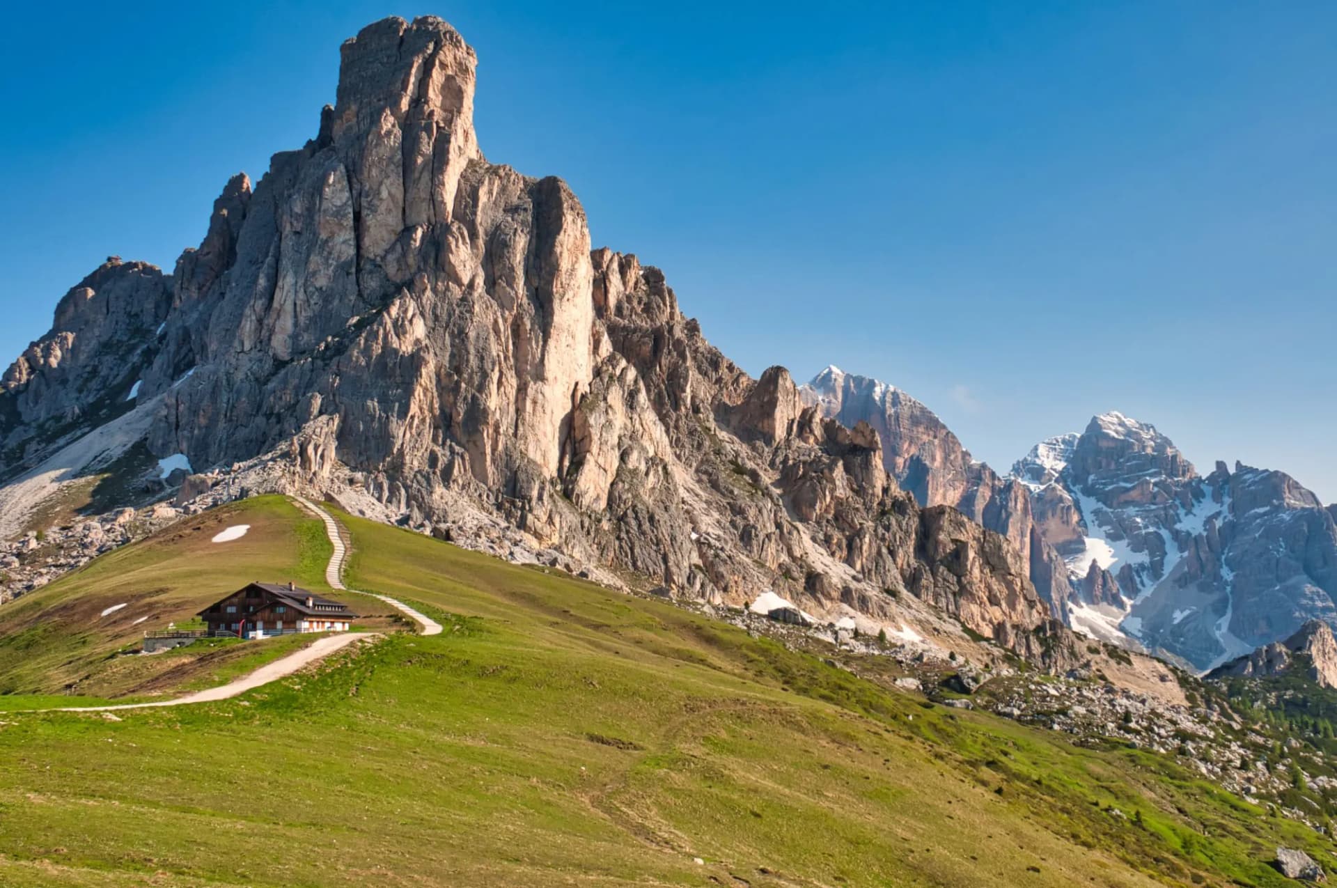 Mountain hut on grassy slope below massive rocky peaks with winding path, Passo Giau.
