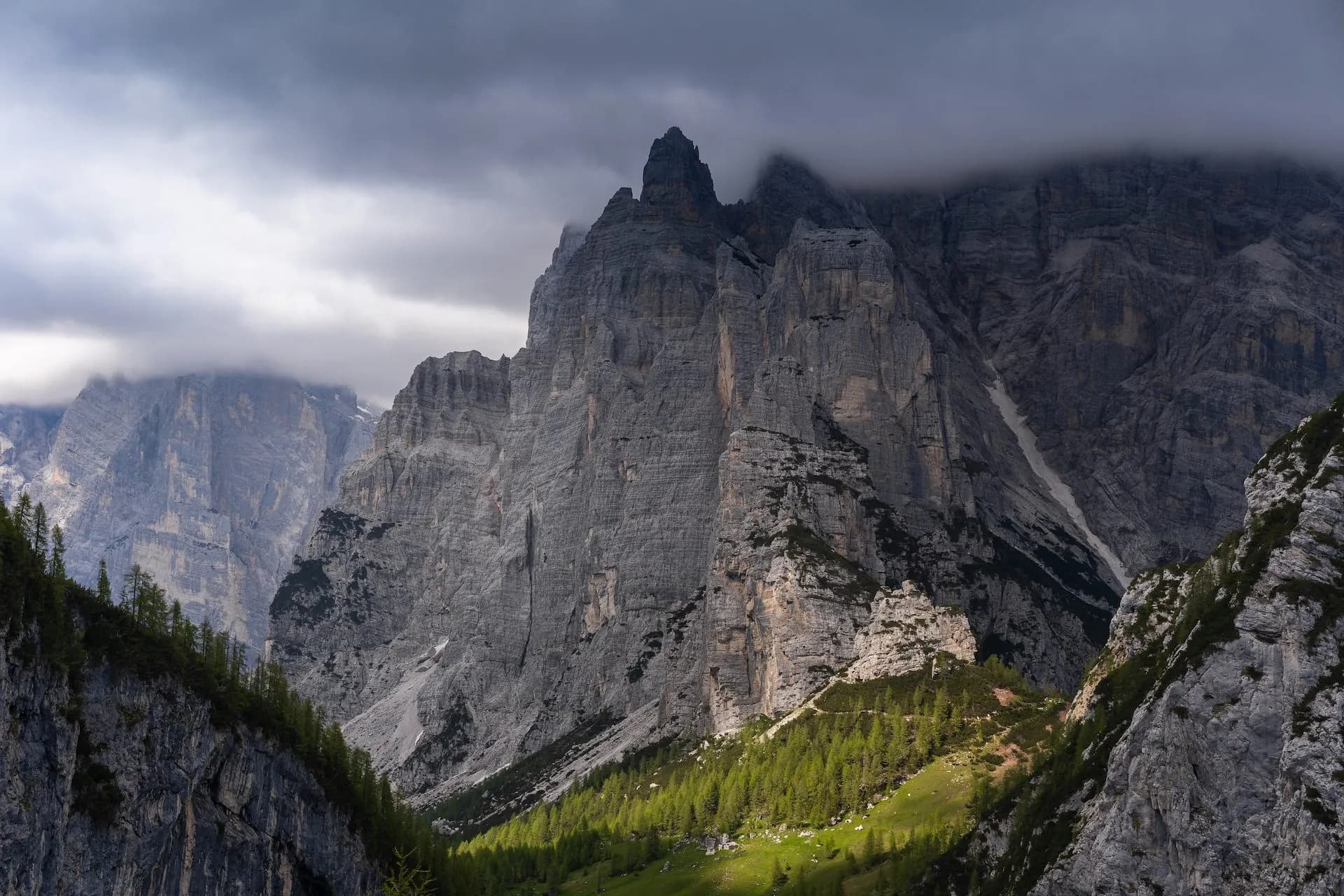 Towering grey rock mountains under dark clouds, with green forest slopes below.