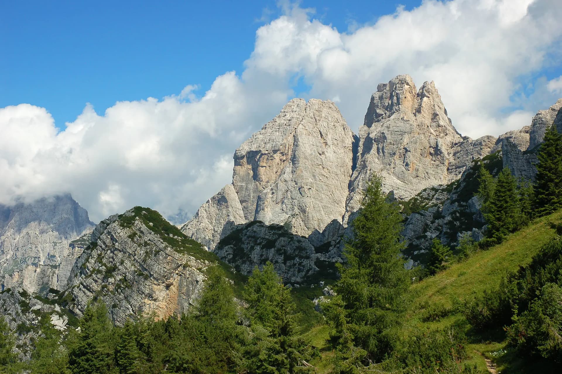Rocky mountain peaks towering over green slopes and pine trees under a blue sky with white clouds.