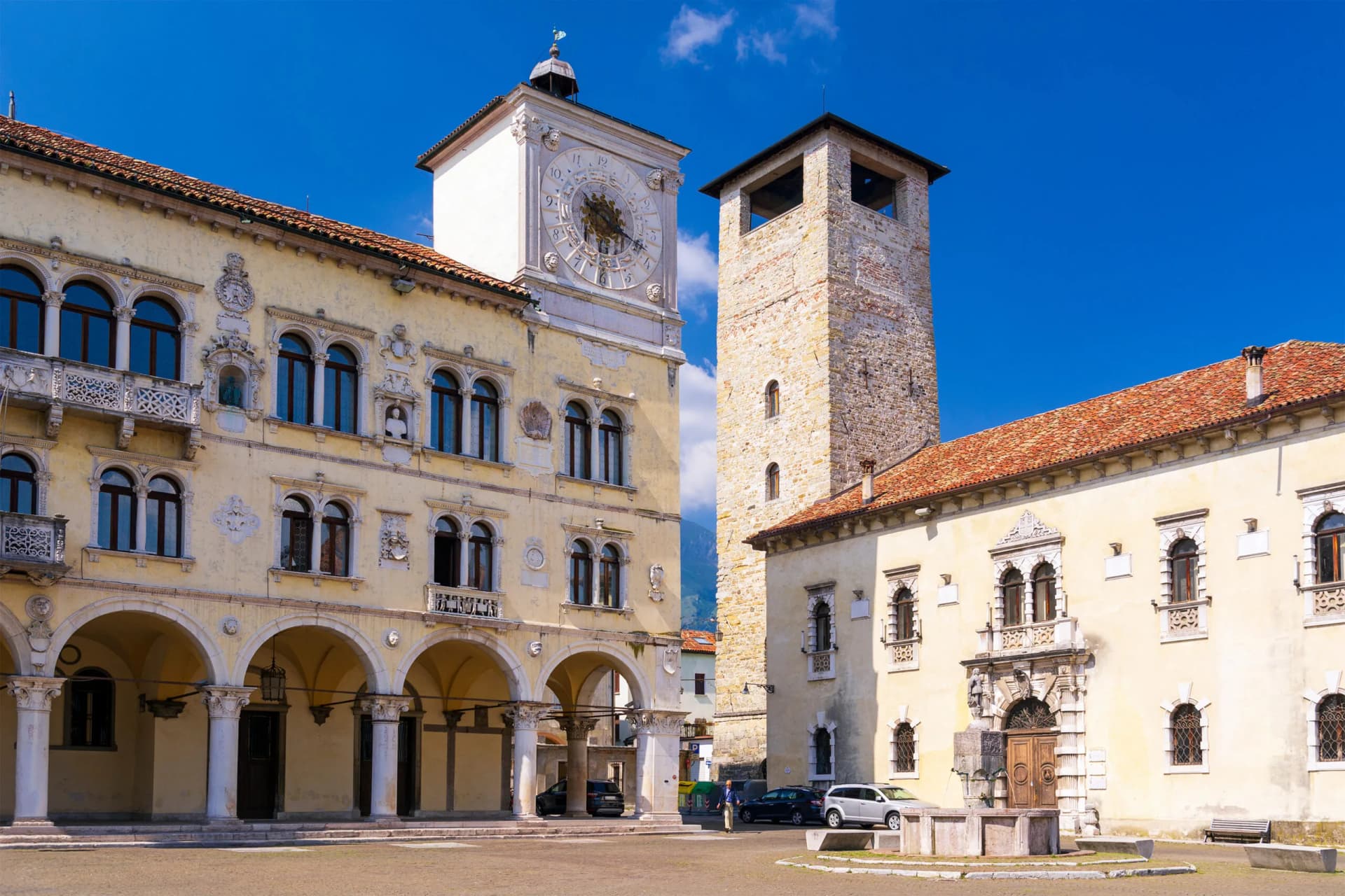 Historic buildings with a clock tower and stone tower in Belluno, Italy under blue sky.