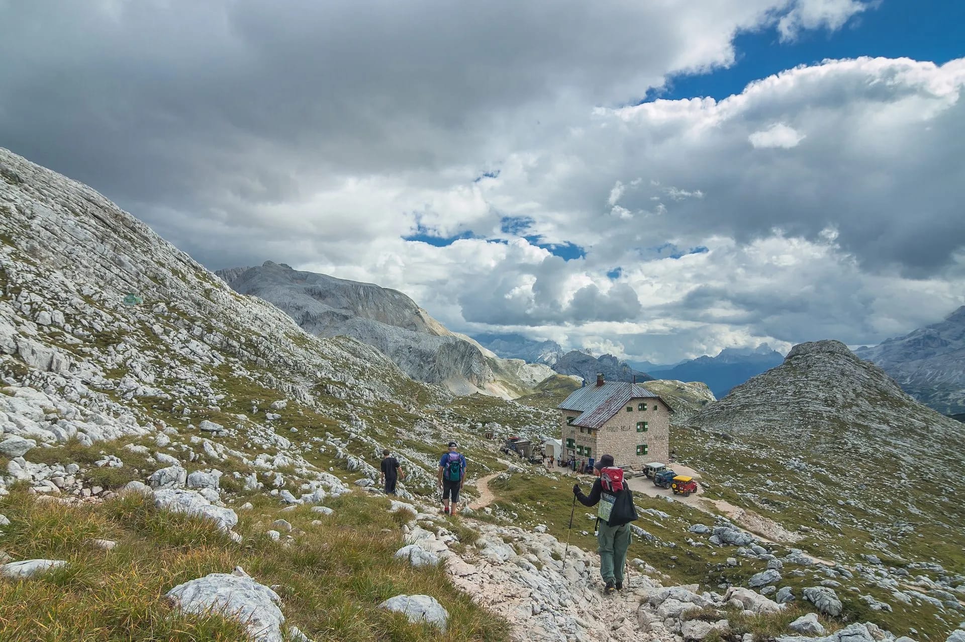 Hikers approaching Rifugio Biella mountain hut among rocky terrain under a dramatic cloudy sky.