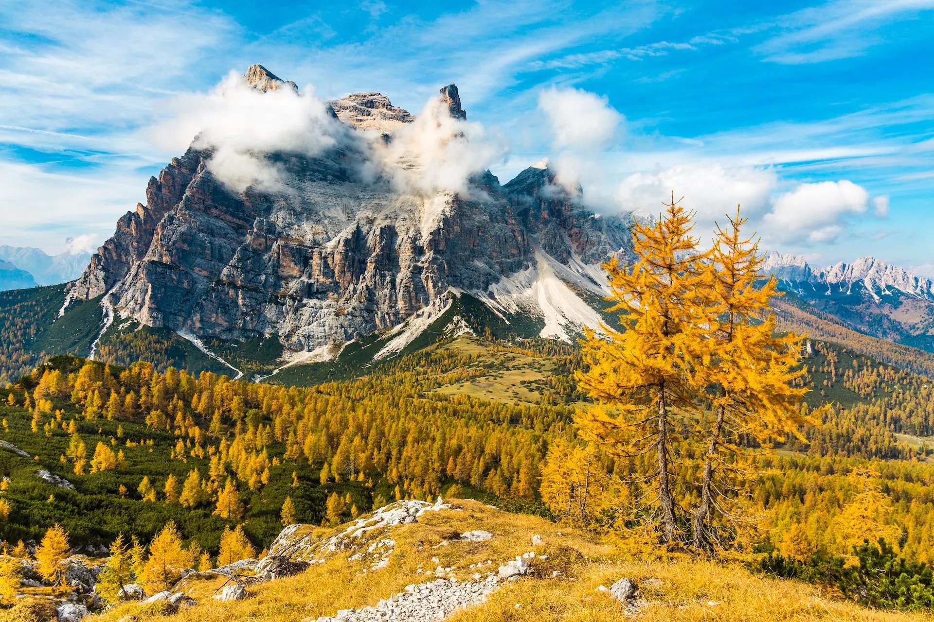 Rugged Monte Pelmo peak with clouds, bright yellow autumn trees, and blue sky.