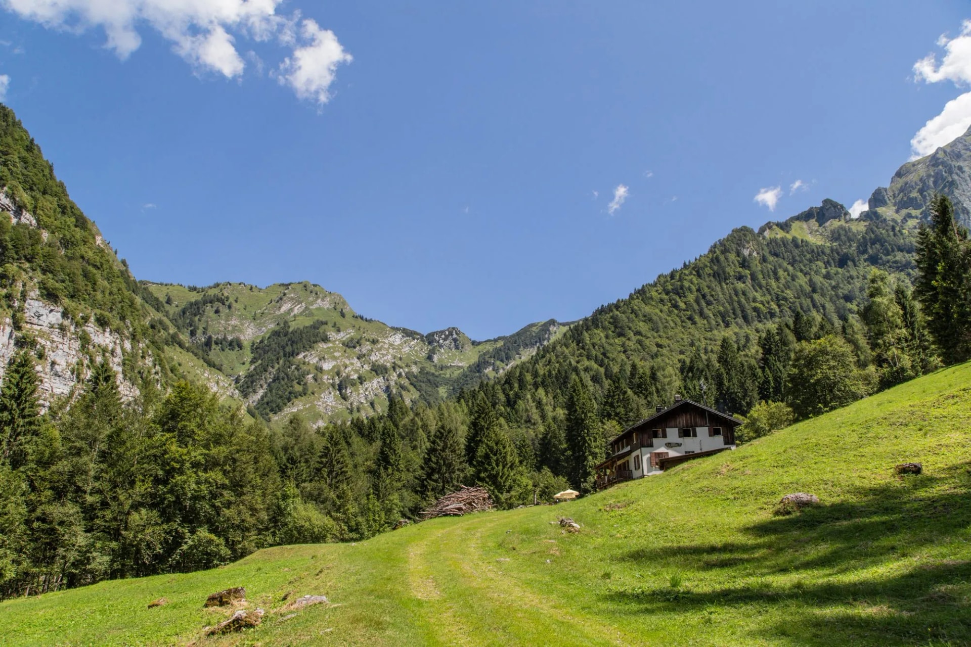 Alpine chalet on grassy slope surrounded by steep, forested mountains under blue sky
