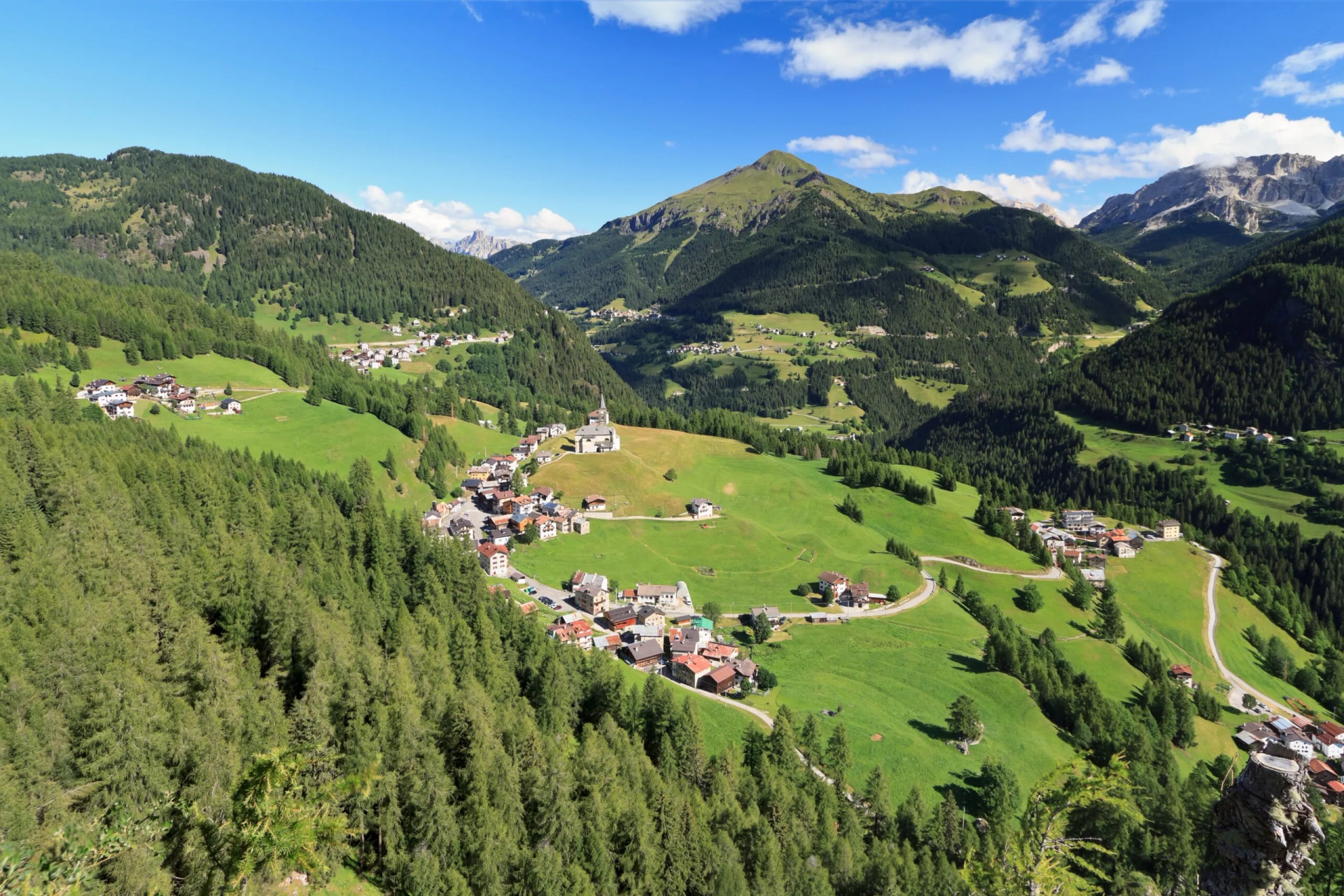 Alpine village nestled in green valley with forested slopes and rocky peaks under blue sky.
