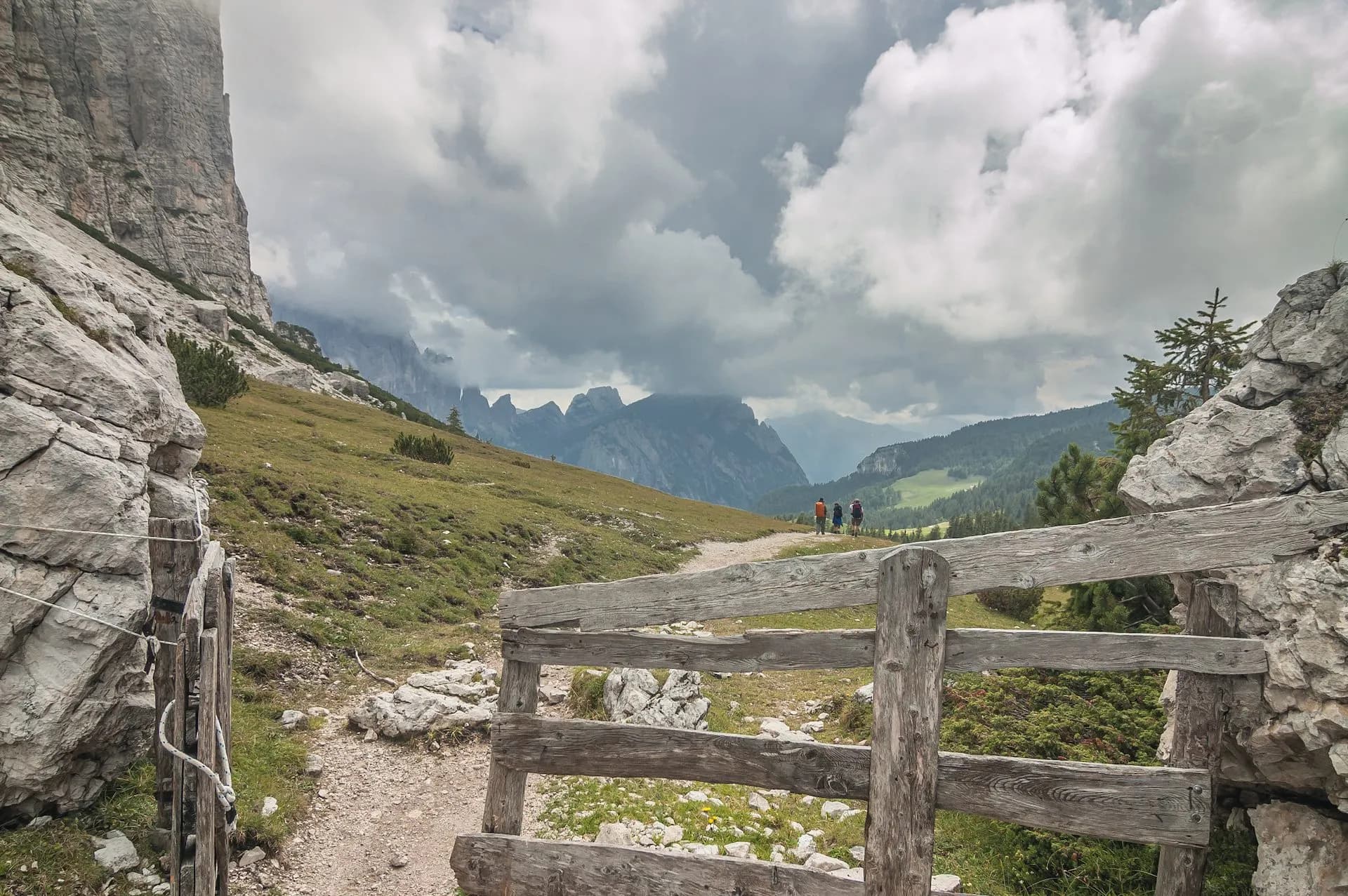 Hikers on dirt path past wooden fence toward jagged mountains under cloudy sky on Alta Via 1.