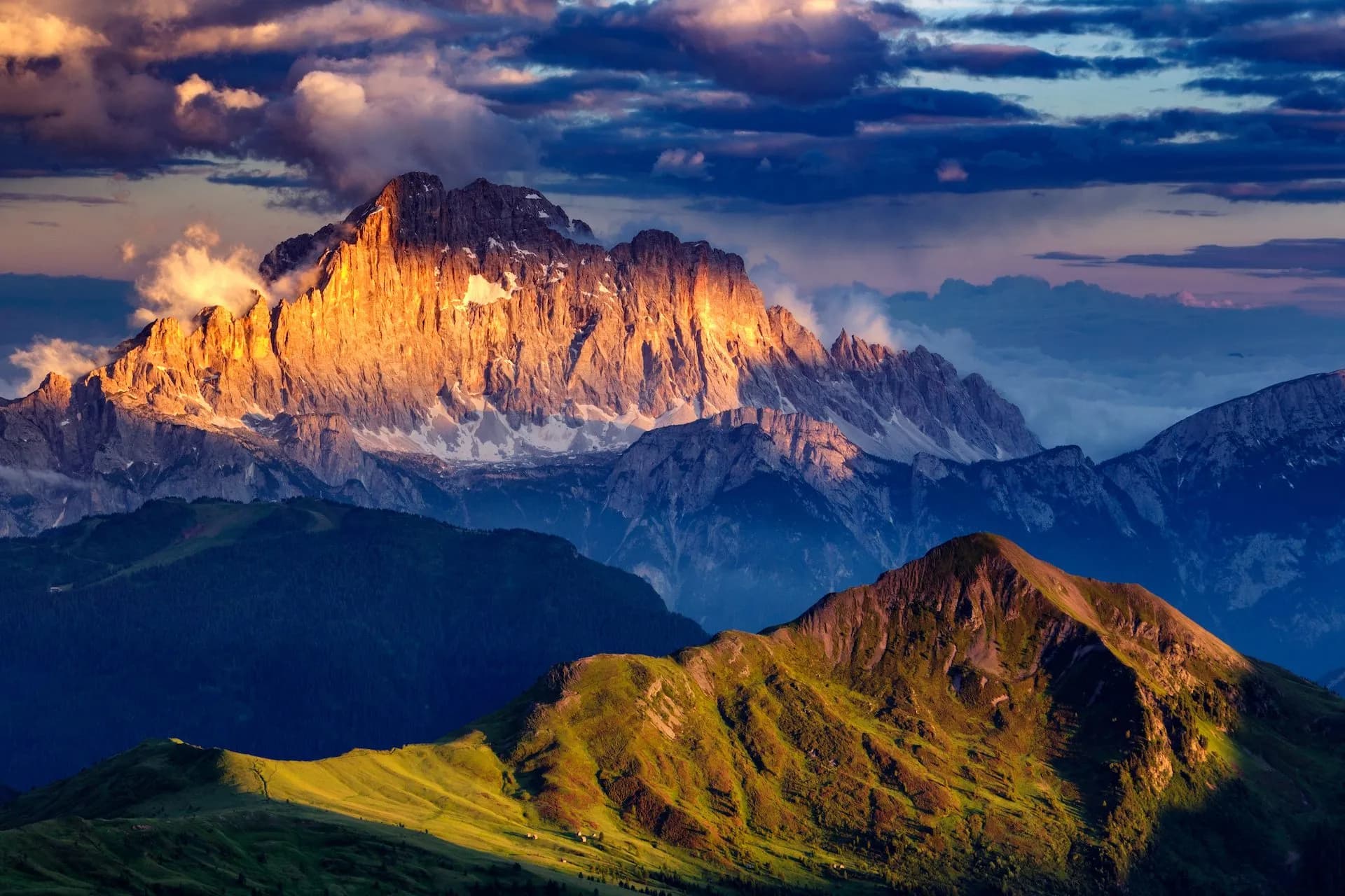 Rugged mountain peaks illuminated by golden sunset light above green foothills under dramatic clouds.