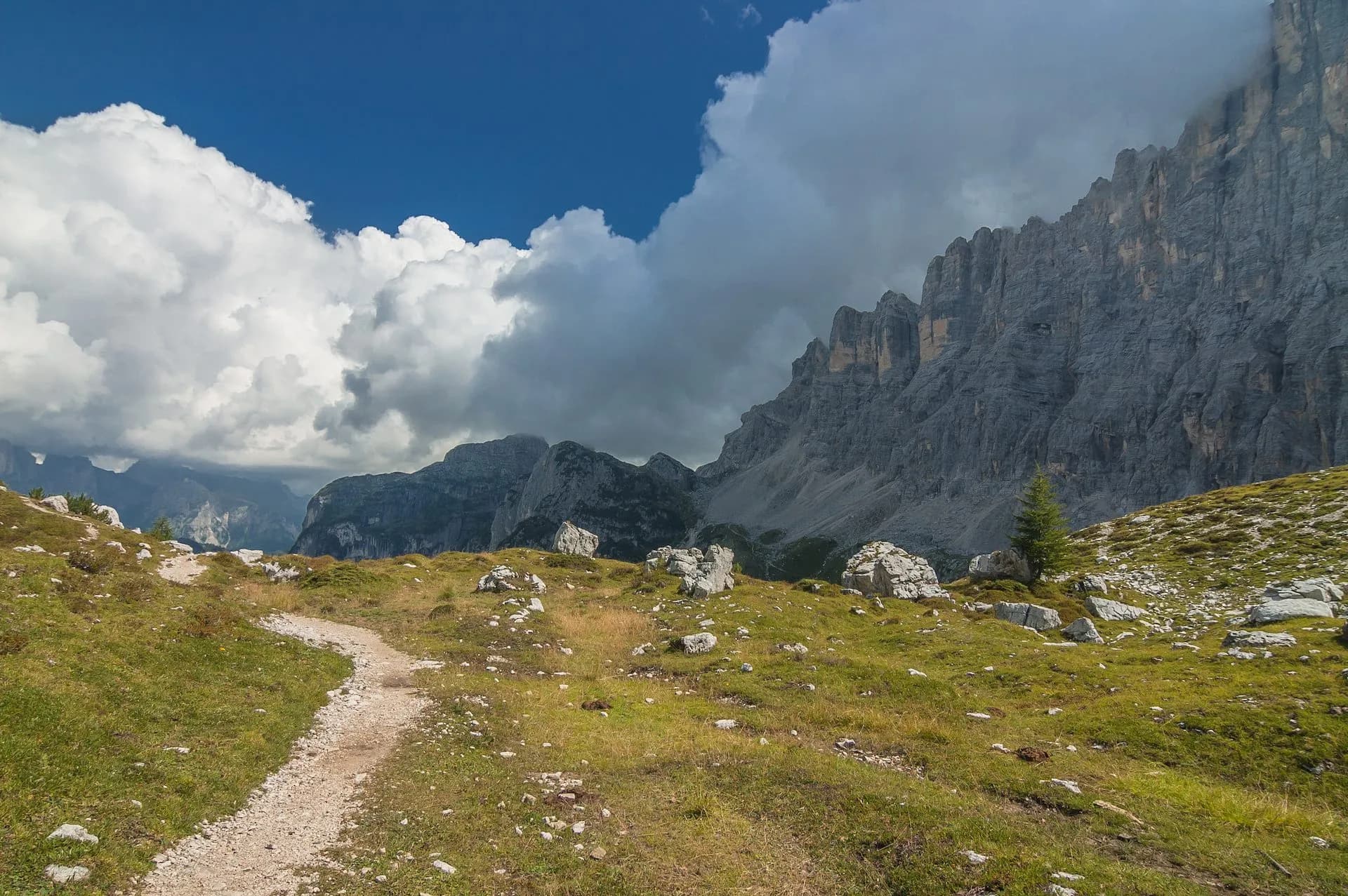 Hiking trail through grassy alpine meadow toward towering gray mountains under dramatic clouds.
