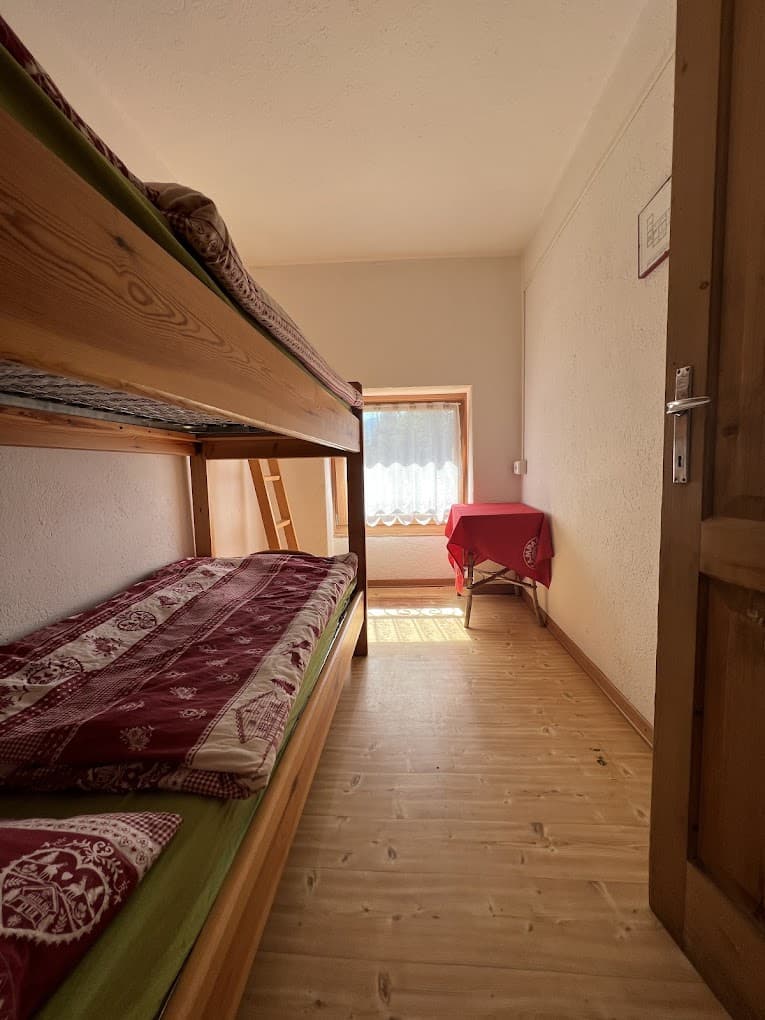 Wooden bunk beds with red patterned bedding in a simple room with light wood floors, Rifugio Vazzoler.