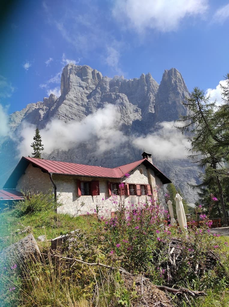 Rifugio Vazzoler stone mountain hut with red roof below jagged peaks and low clouds.