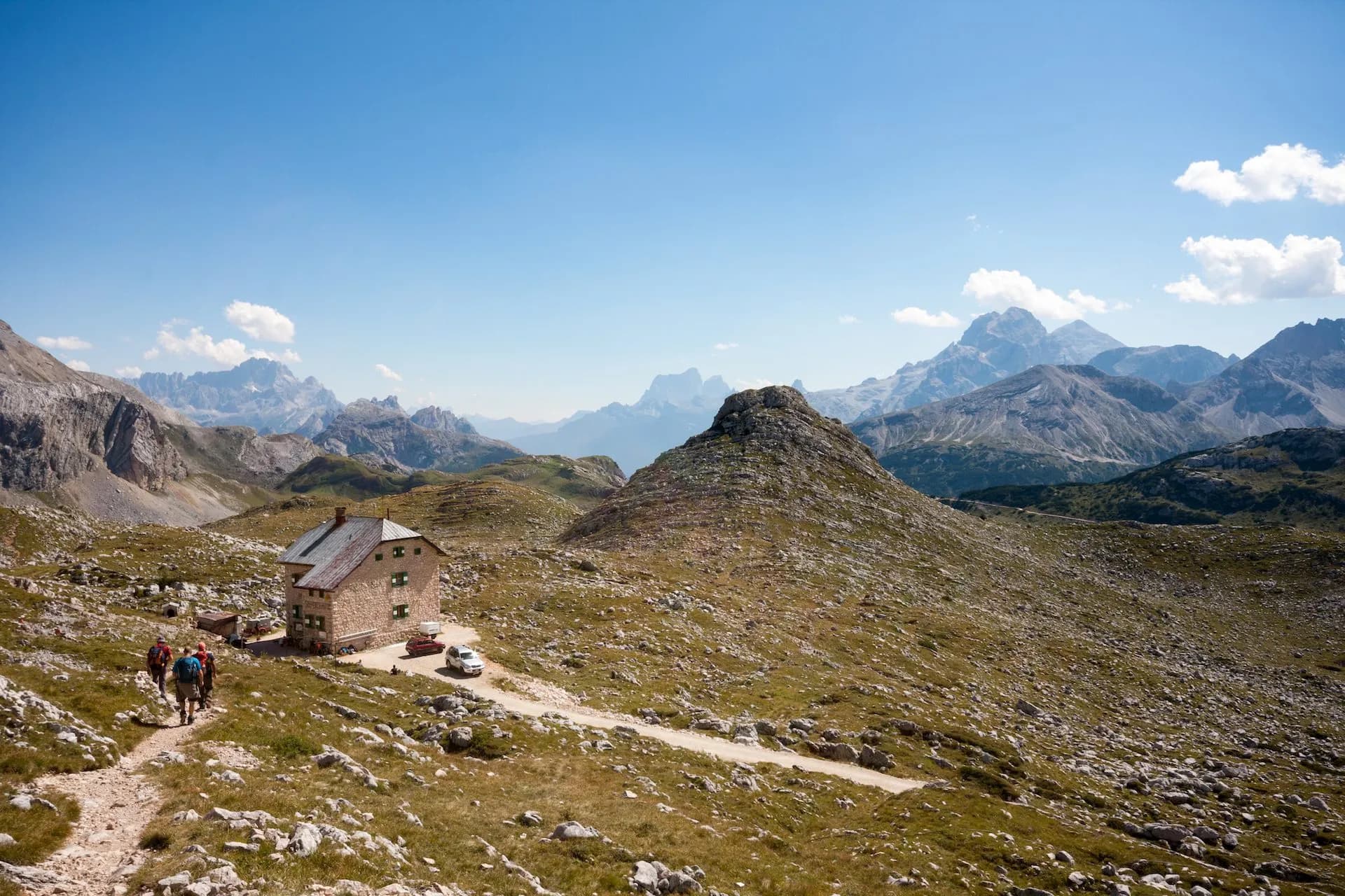 Hikers approach Rifugio Biella stone mountain hut surrounded by rocky alpine terrain and distant peaks.