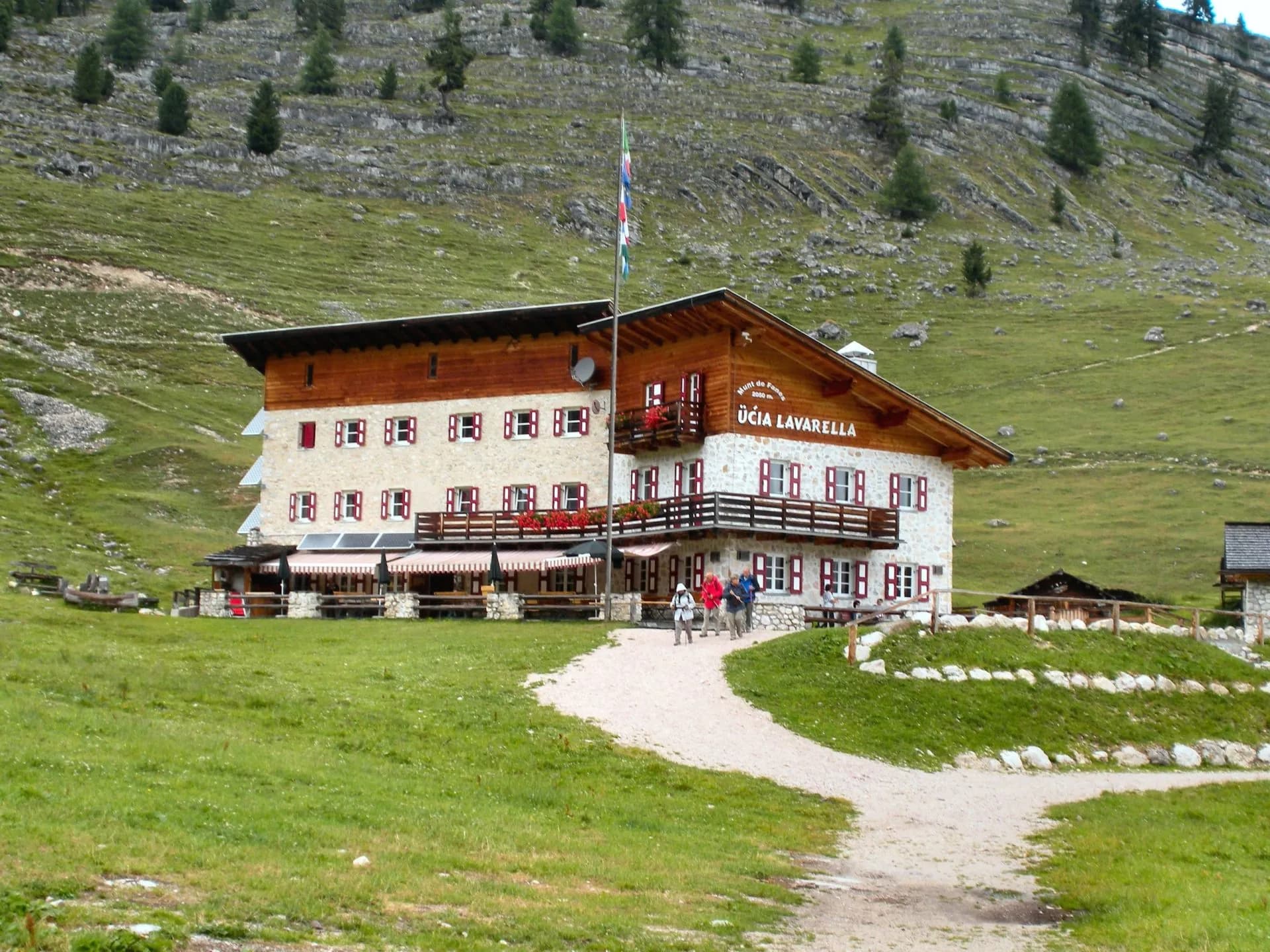 Rifugio Lavarella mountain hut with hikers approaching on a dirt path below grassy slopes.