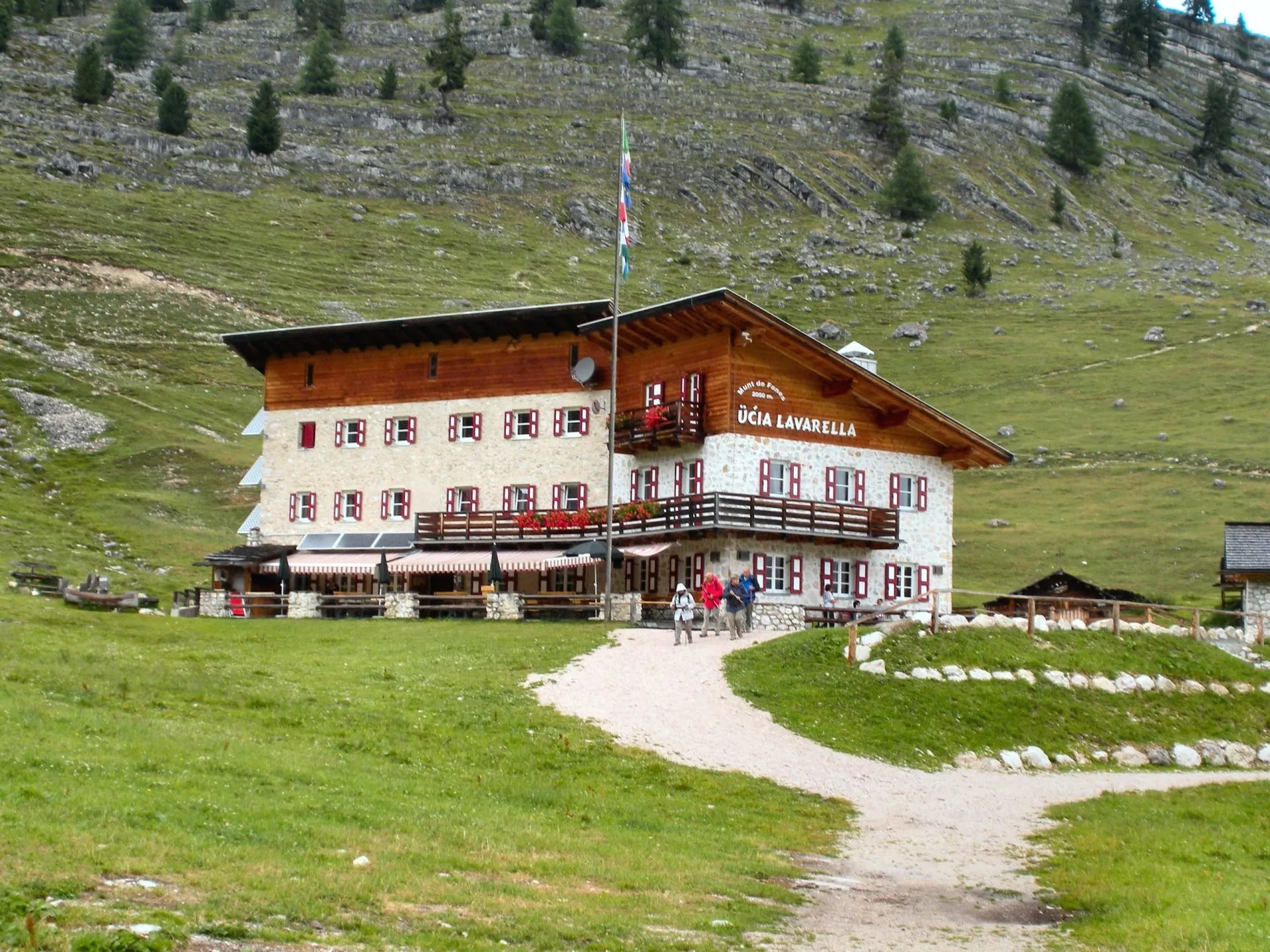 Rifugio Lavarella mountain hut with hikers approaching on a dirt path below grassy slopes.