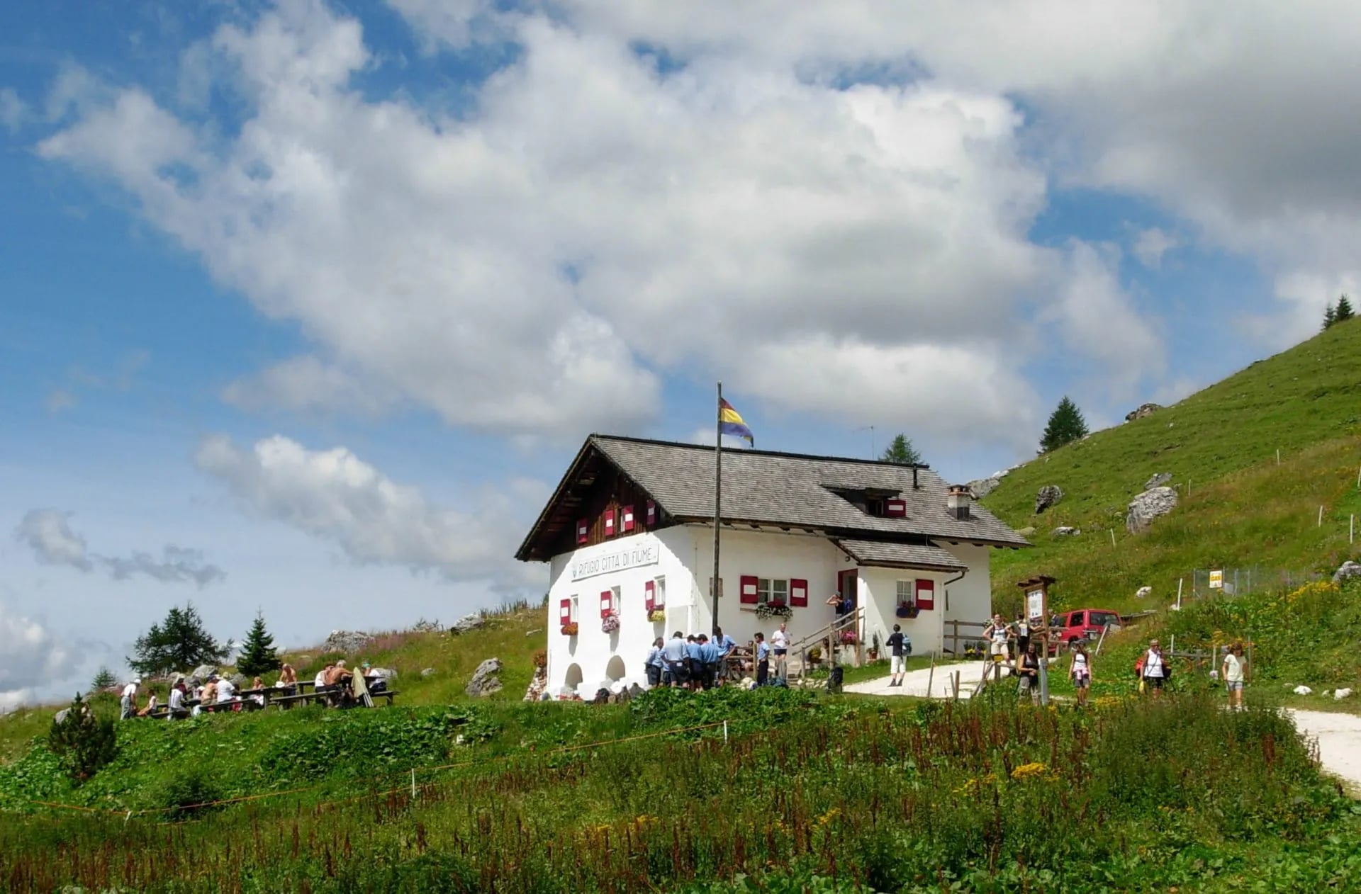Rifugio Città di Fiume mountain hut with hikers on a grassy slope under a cloudy sky.