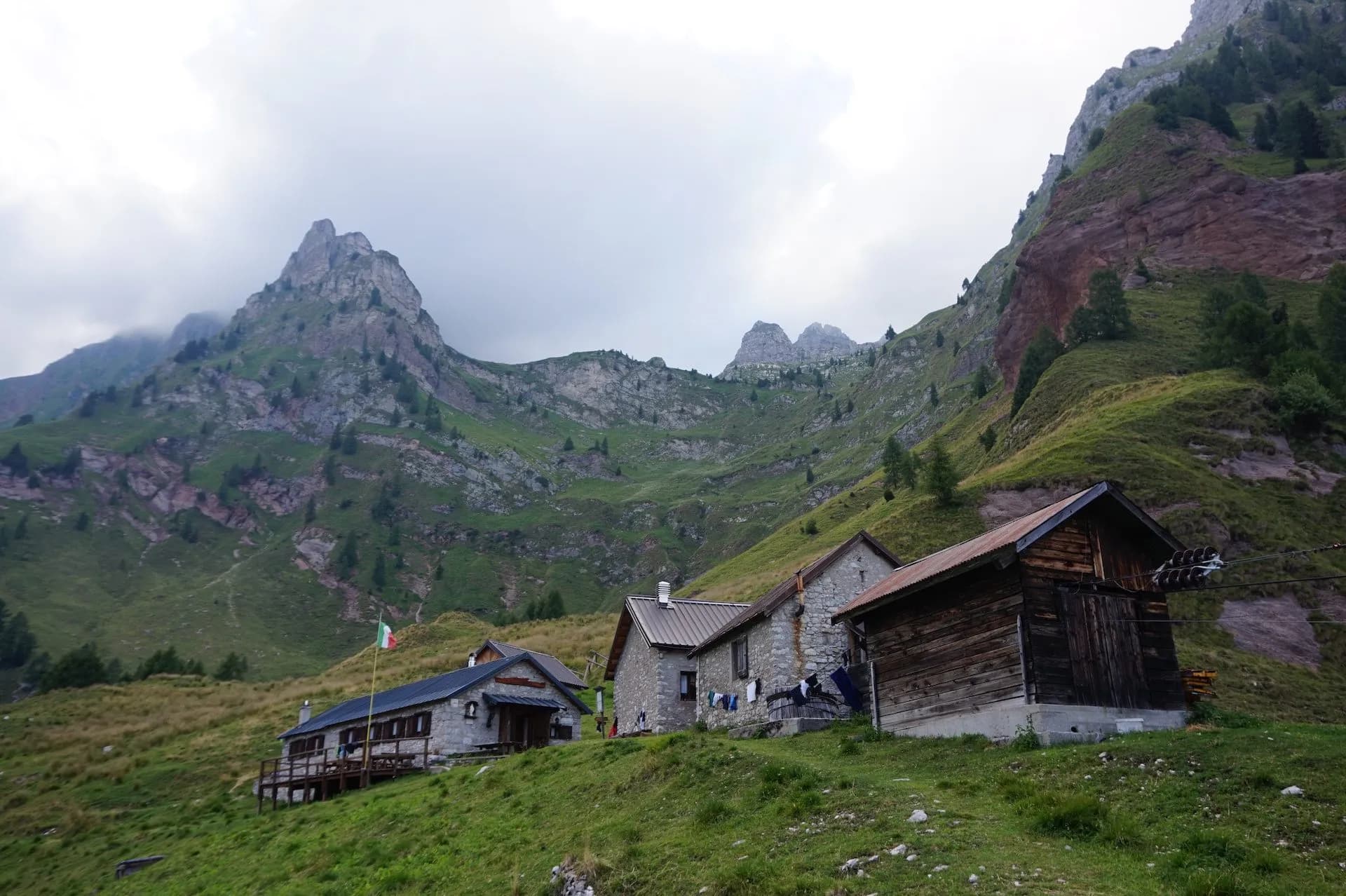 Rifugio Pian de Fontana stone huts nestled on a grassy slope beneath rugged green mountains.