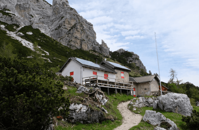Mountain hut with white walls and red shutters nestled below steep rocky slopes and green vegetation.