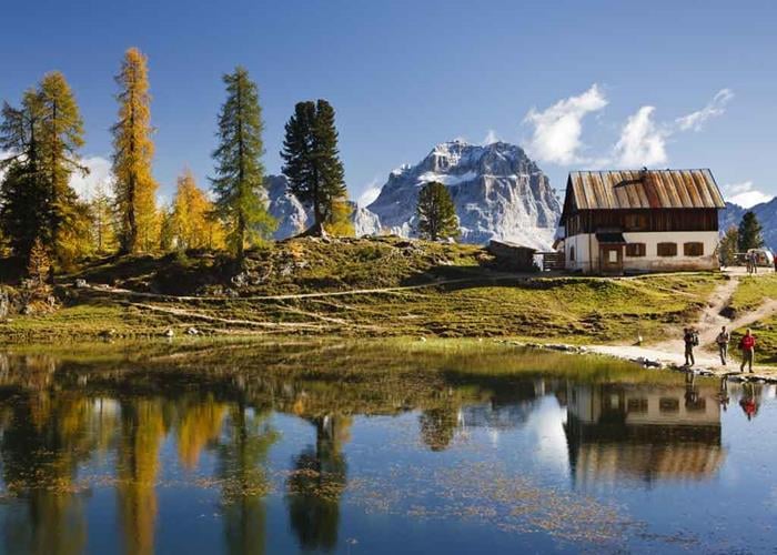Rifugio Croda da Lago hut by mountain lake with hikers and autumn trees.
