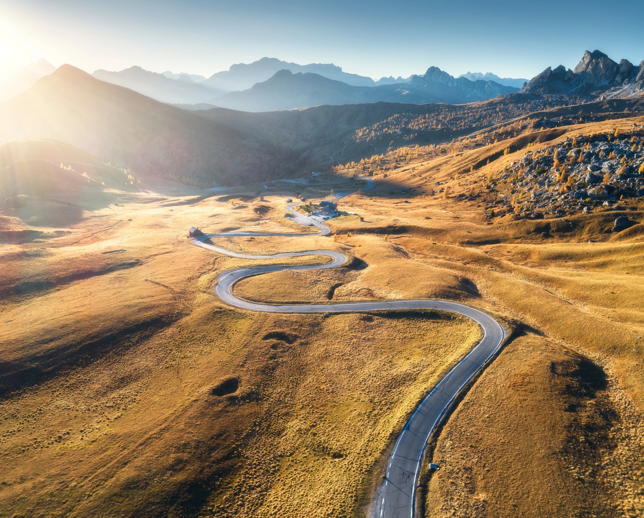 Winding road in mountain valley at sunset in autumn. Aerial view of asphalt road in Passo Giau. Dolomites, Italy. Top view of roadway, mountains, meadows with orange grass, blue sky and gold sunlight