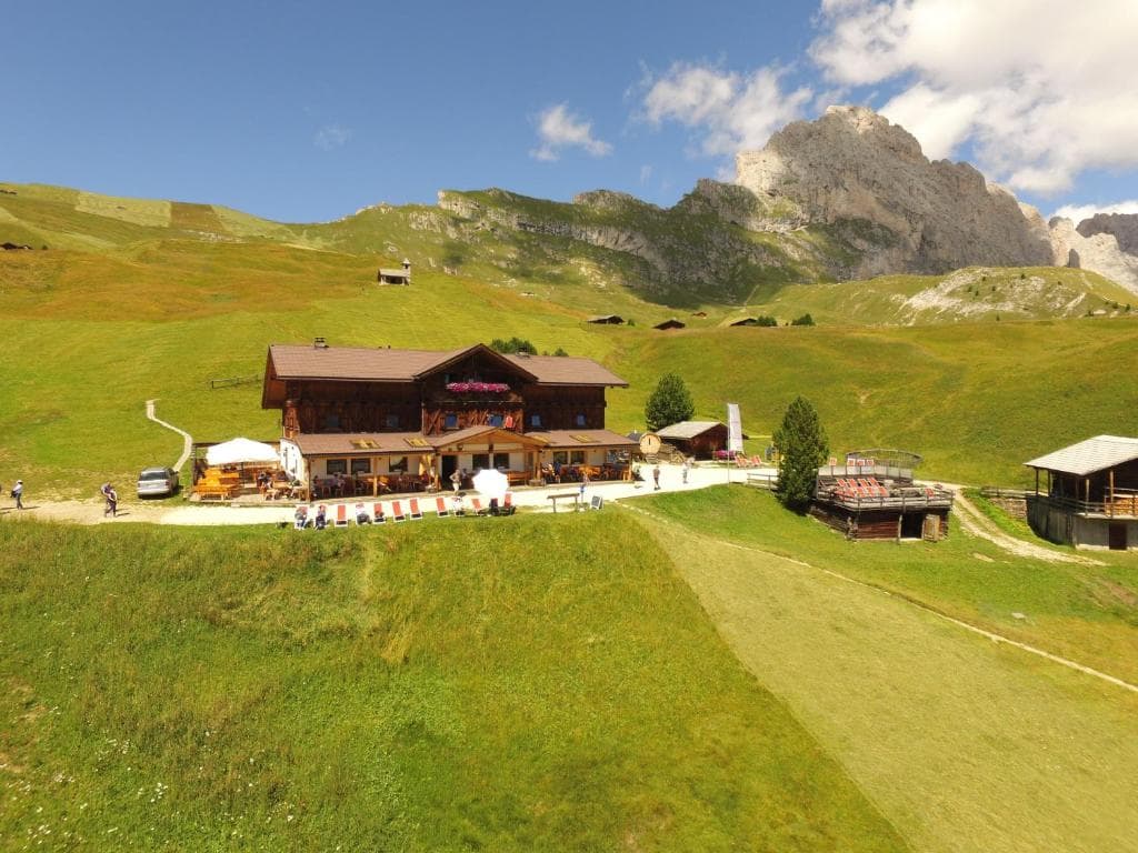 Alpine mountain lodge with outdoor dining area in a grassy valley with rocky peaks.