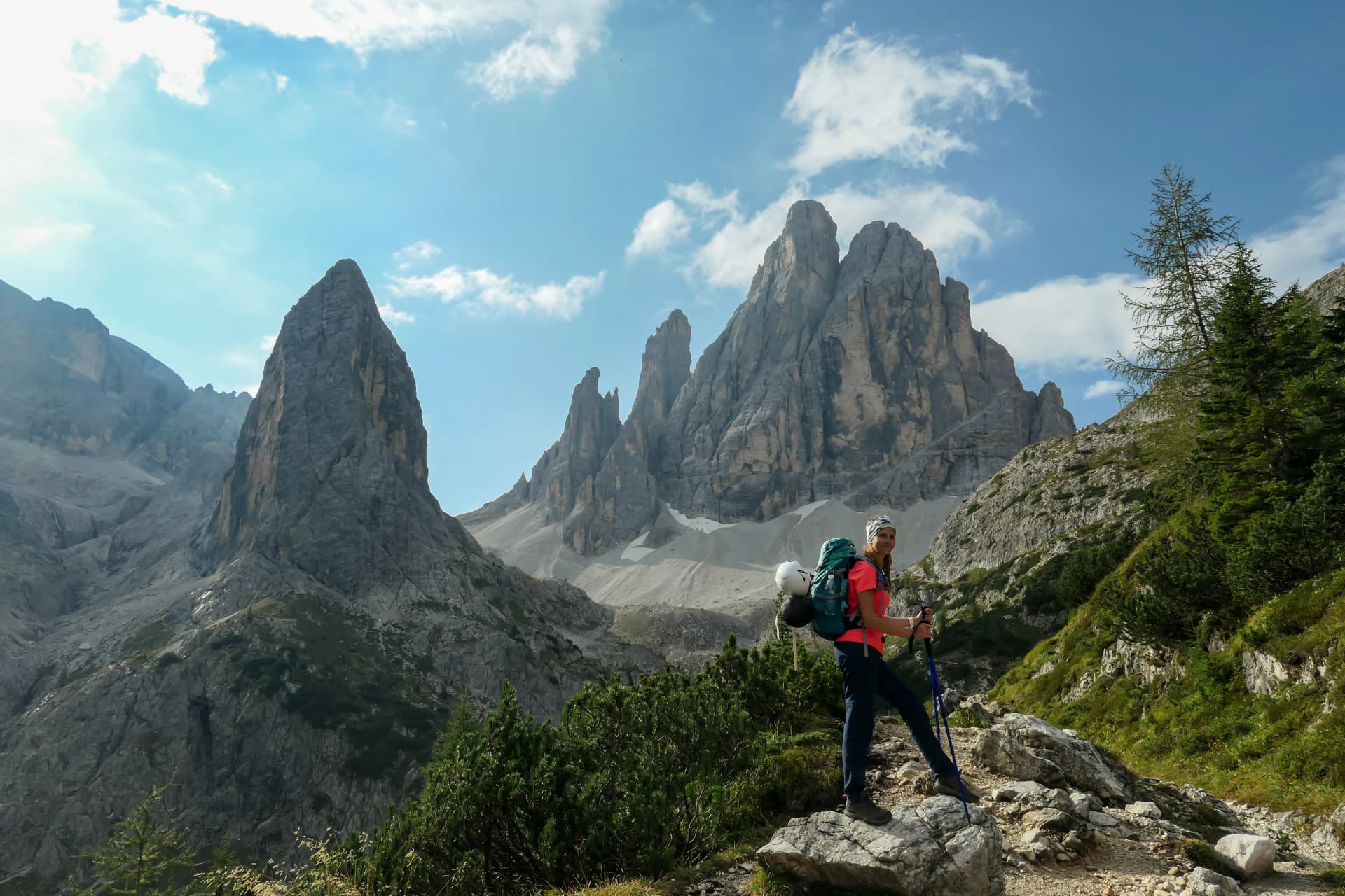 A woman with big hiking backpack hiking in high Italian Dolomites. There are many sharp peaks behind her. She is enjoying the hike, smiling. There are a few trees around. Sunny day. Outdoor exercising