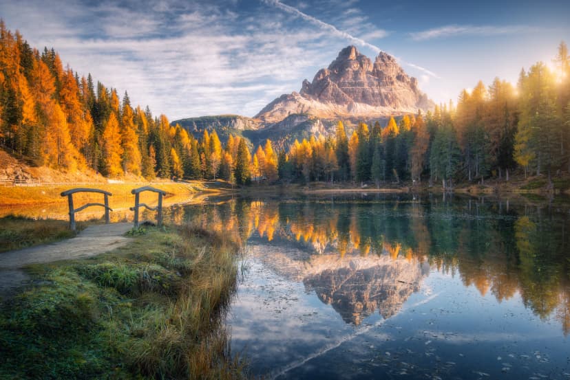 Lake with reflection in mountains at sunrise in autumn in Dolomites, Italy. Landscape with Antorno lake, small wooden bridge, trees with orange leaves, high rocks, blue sky in fall. Colorful forest