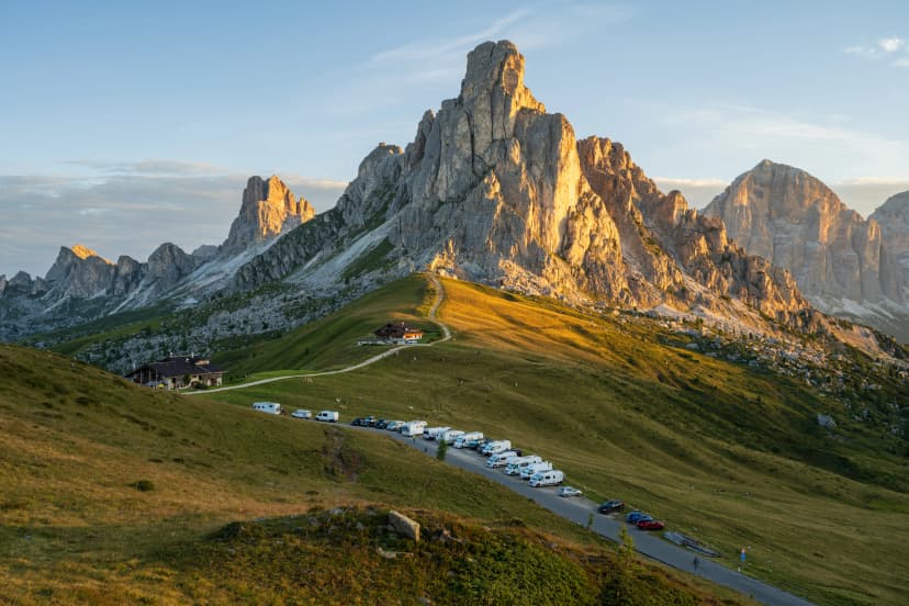 Sunrise at Passo Giau with road and parked cars and campers with sun shining at meadow and peaks in the background during summer morning in august