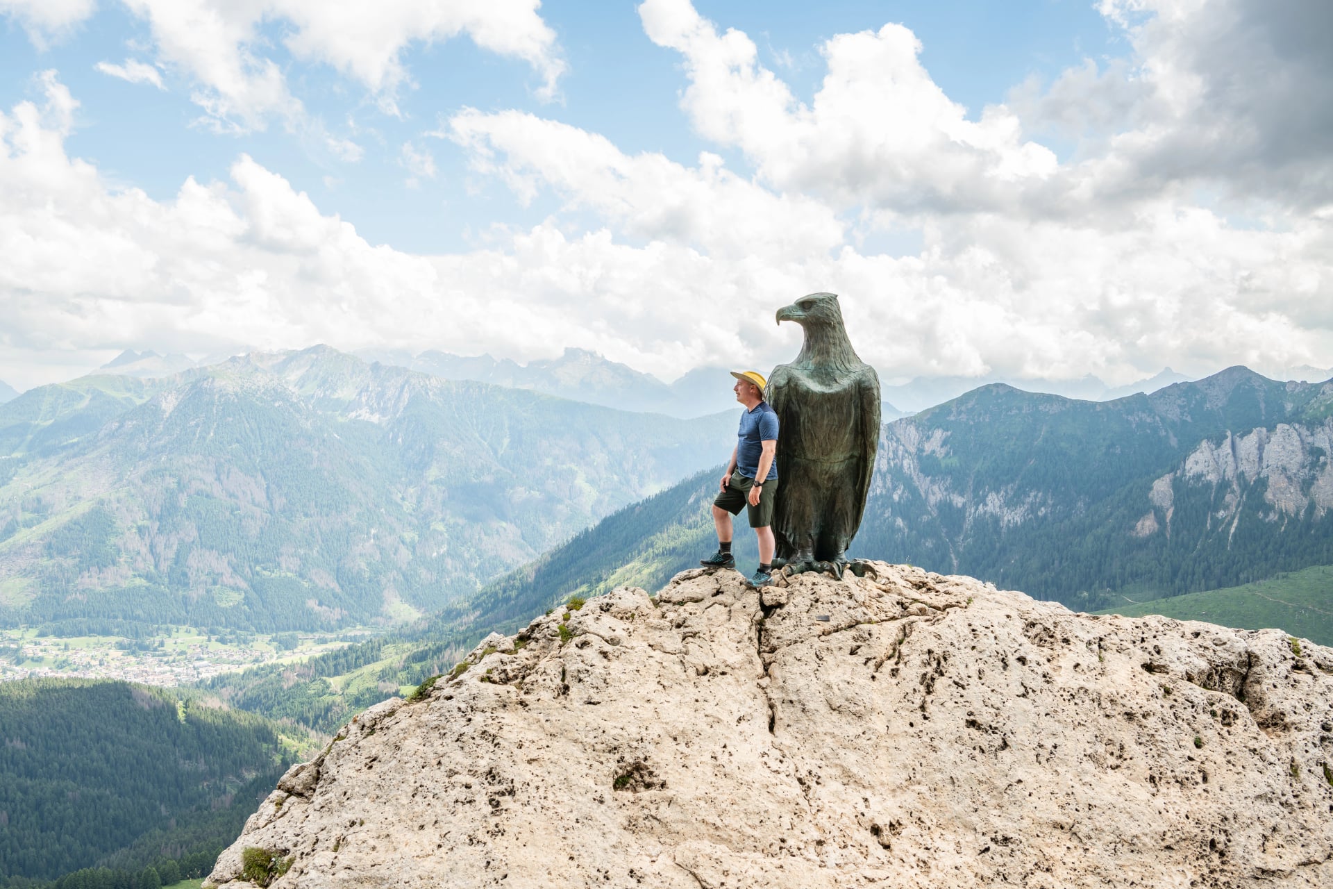 13 july 2024, Roda di Vael, Italy. Caucasian hiker standing at the Christomannos monument, a bronze eagle in front of the mighty walls of the Catinaccio-Rosengarten massif Italian Dolomites, hiking d