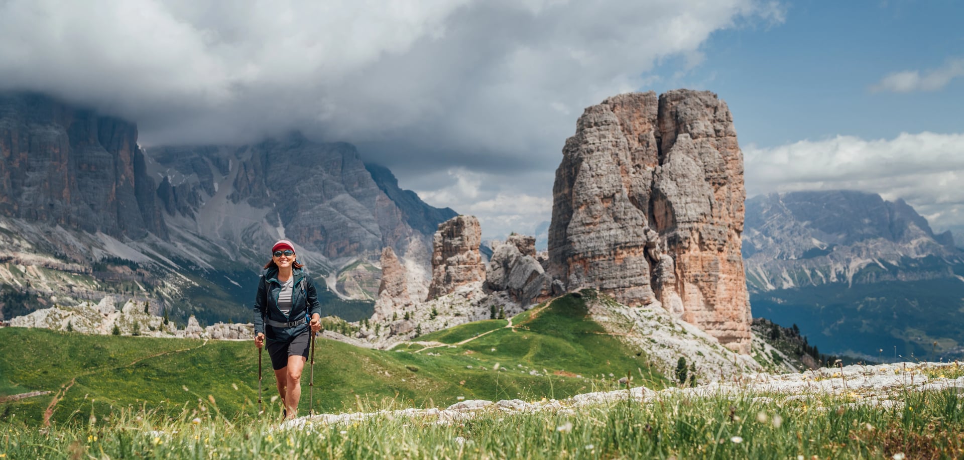 Smiling female trekker walking with backpack and trekking poles by green mountain hill with picturesque Dolomite Alps Cinque Torri formation on the background. Active people and mountains concept.