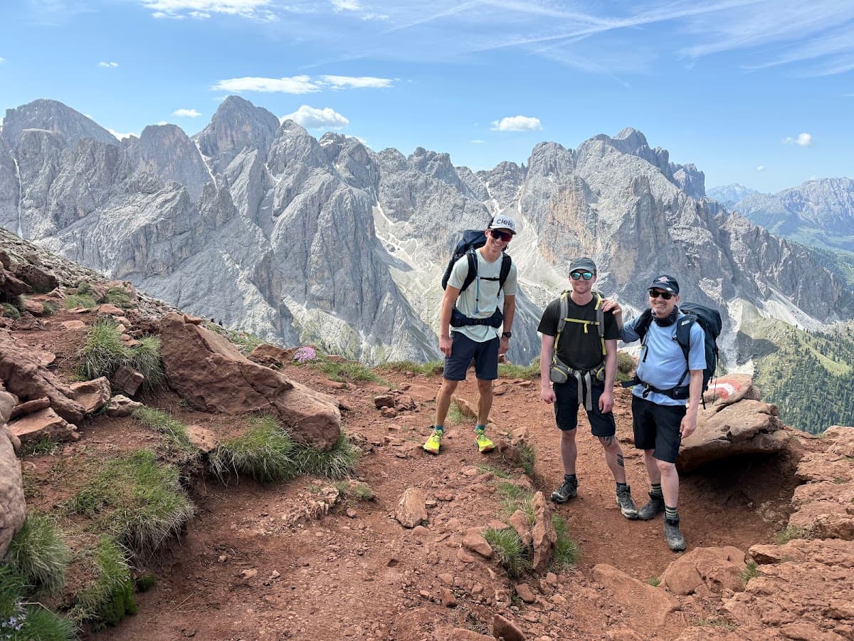 Three hikers with backpacks standing on a rocky mountain trail with alpine peaks