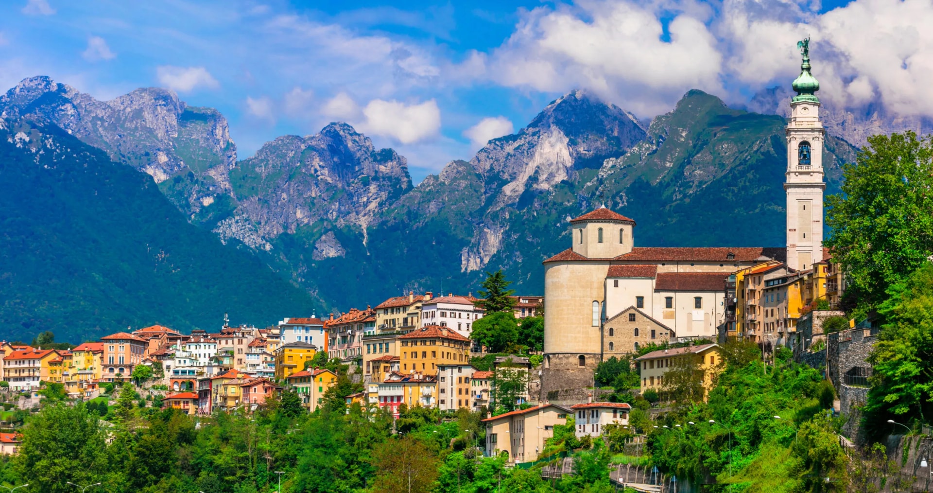 Belluno town with colorful buildings and church tower nestled below impressive green and rocky Dolomite mountains.