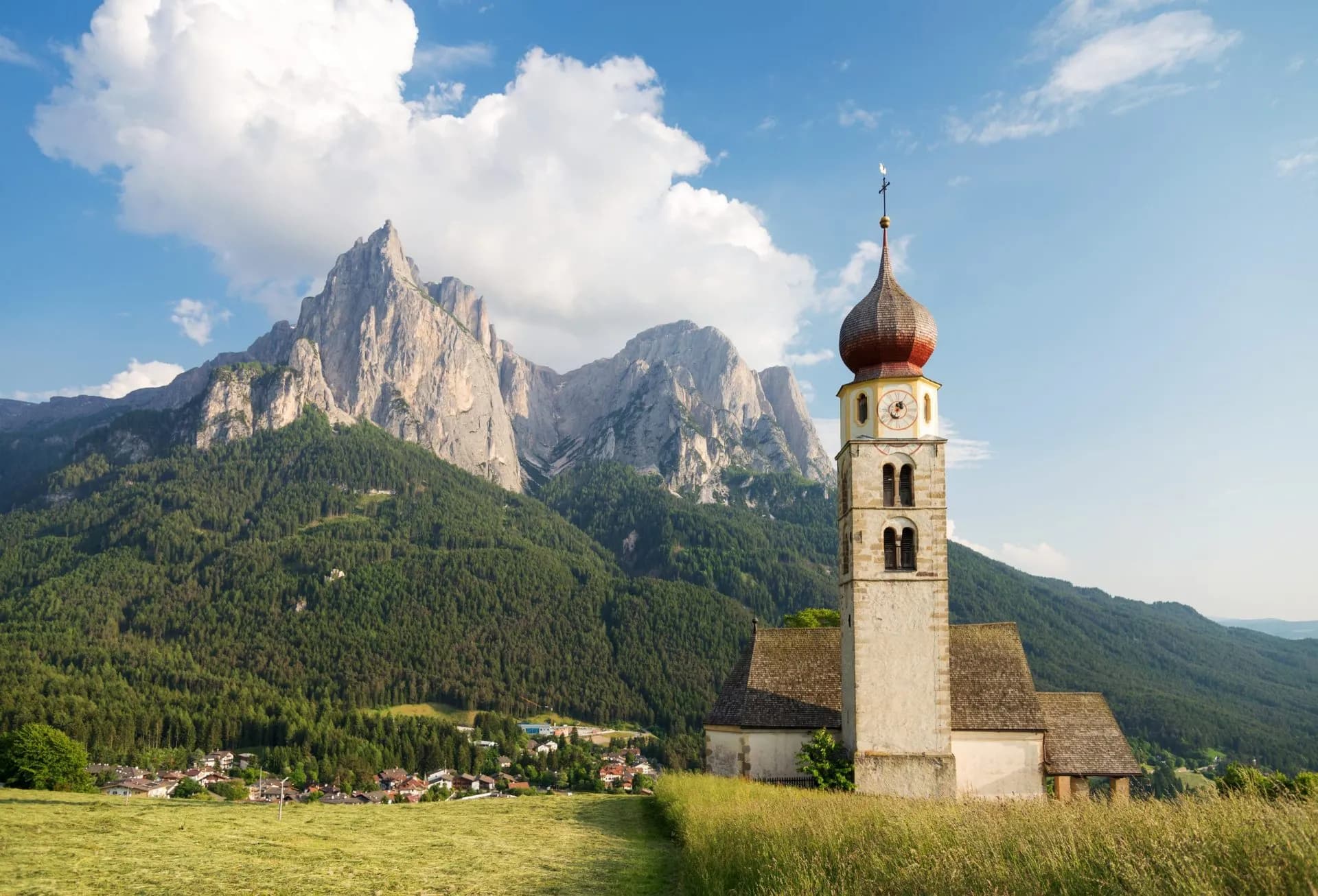 Church with onion dome tower in field before massive rocky mountains and village, Siusi allo Schlern.