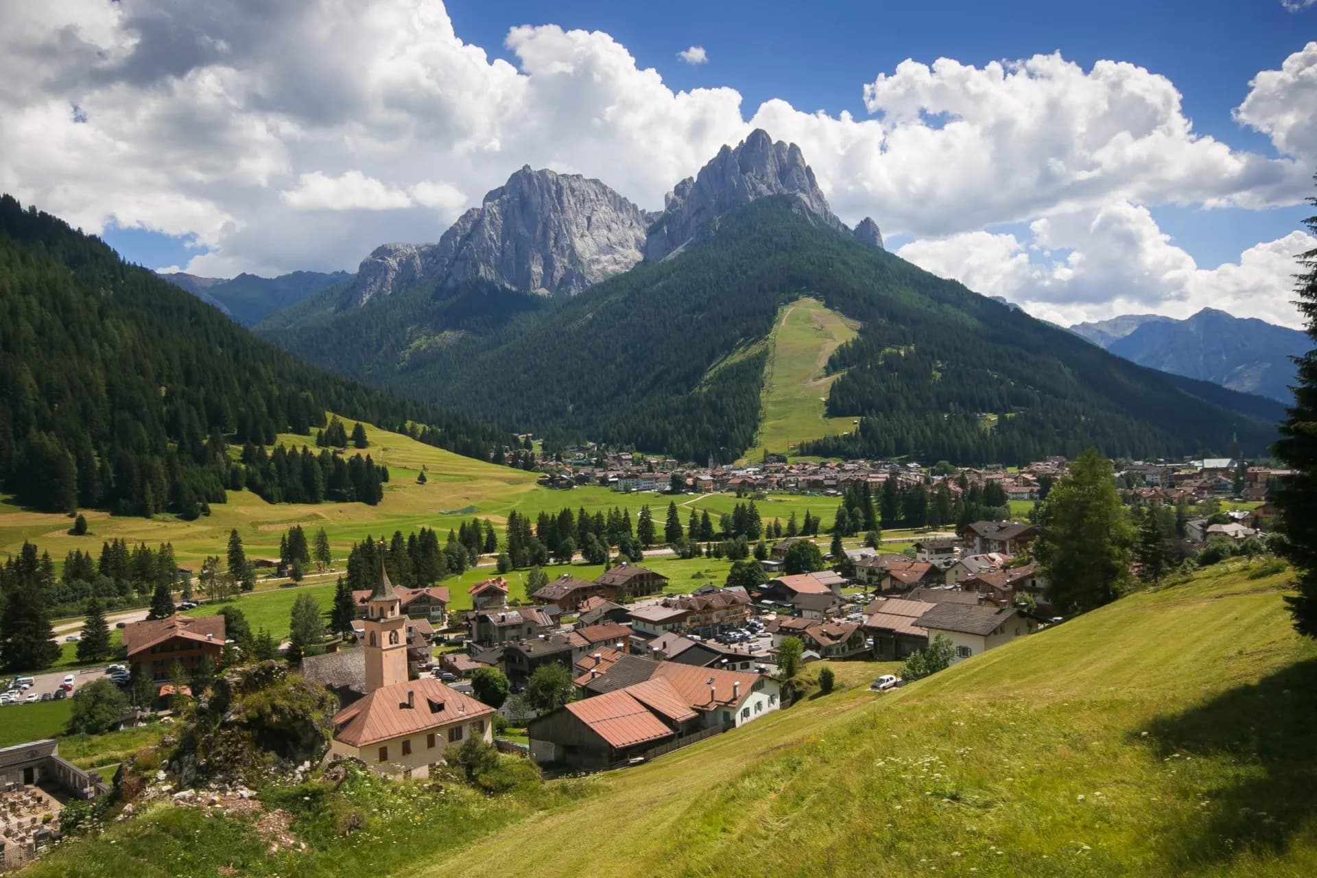 Alpine village nestled in a valley with dense forests and jagged peaks under a cloudy blue sky.