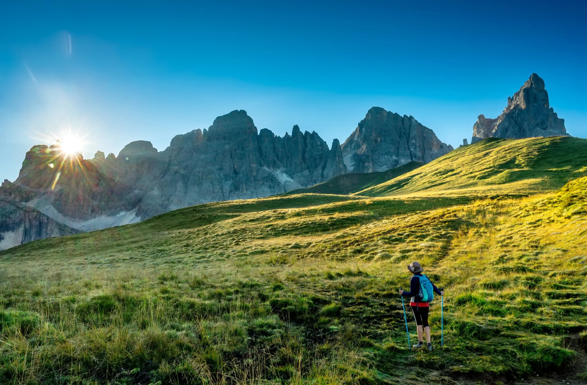 Hiker with trekking poles on grassy slope below jagged mountain range at sunrise, Pale di San Martino.