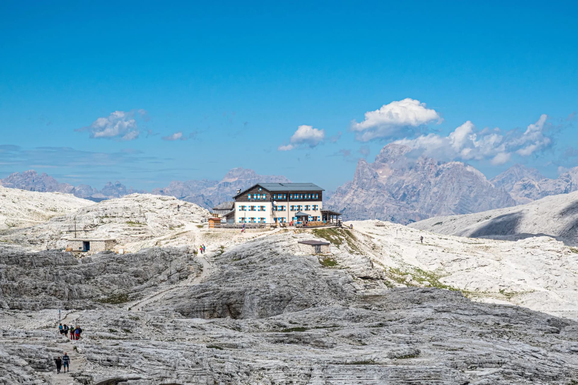 Rifugio Rosetta mountain hut on a rocky plateau with hikers and rugged peaks under a blue sky.