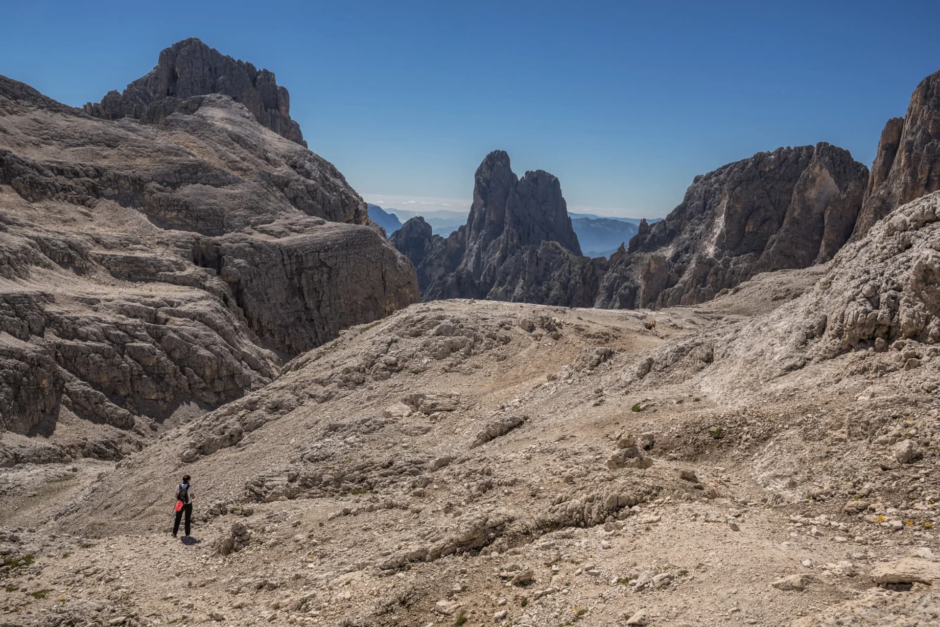 Hiker on rocky, barren terrain surrounded by steep, jagged mountains under a clear blue sky.