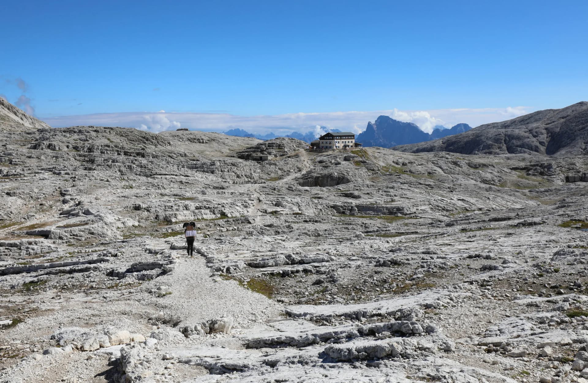 Hiker walking toward Rifugio Pale di San Martino across rocky alpine terrain.