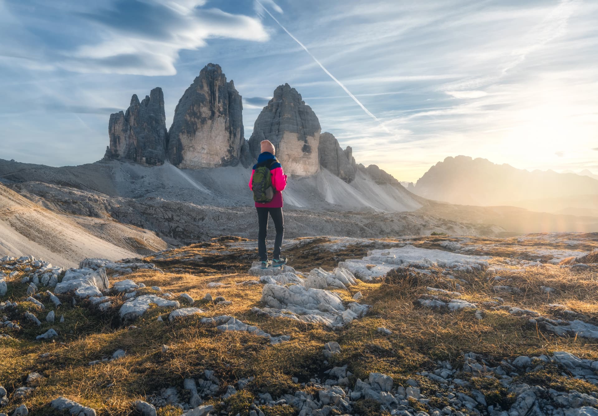Hiker on trail near Tre Cime di Lavaredo mountains at sunset with bright sky.