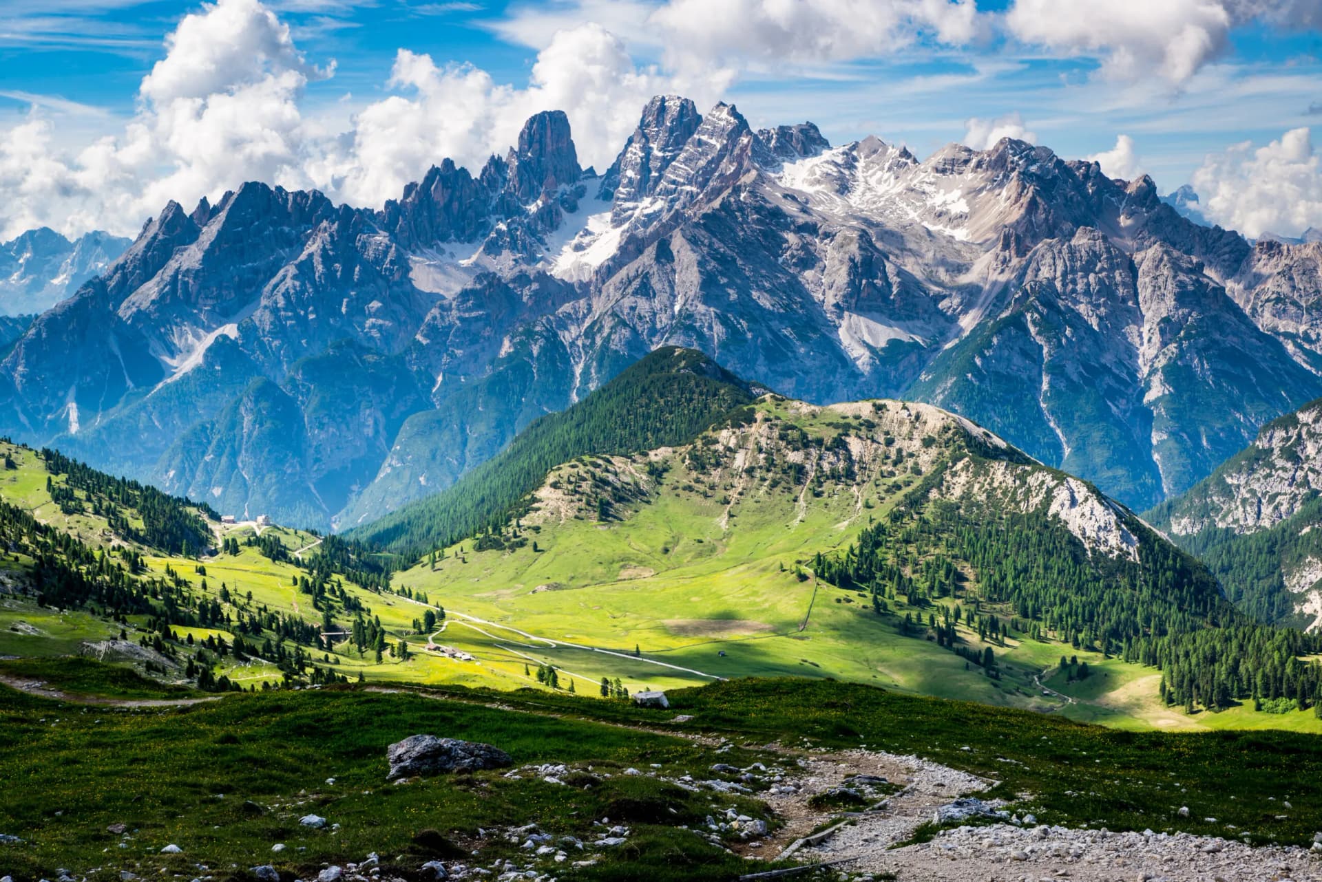 Mountain landscape with snow-capped peaks, green alpine meadows, and a rocky hiking trail.