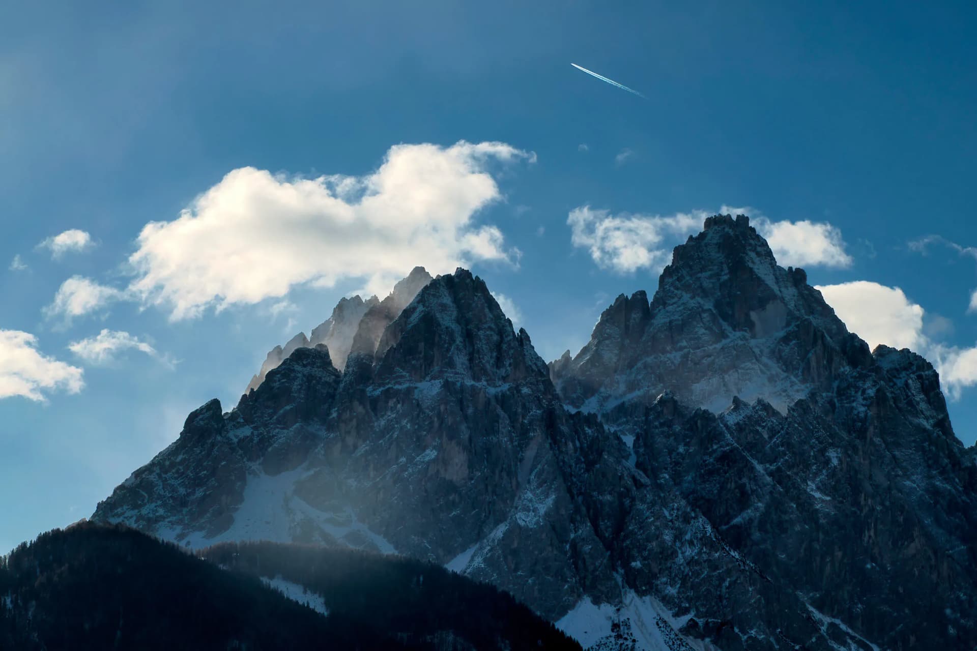Jagged, snow-dusted Dolomites mountains under a blue sky with clouds and a jet contrail, viewed from Dobbiaco.